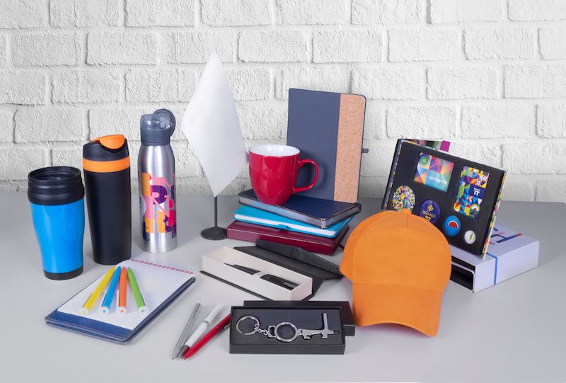 Blank promotional incentive items available at Kevins Worldwide, including. a hat, a bottle opener, three pens, a notebook with highlighters, a coffee mug, a water bottle, a stainless steel water bottle, a small desktop flag, a coffee mug and folder on top of a stack of notebooks.