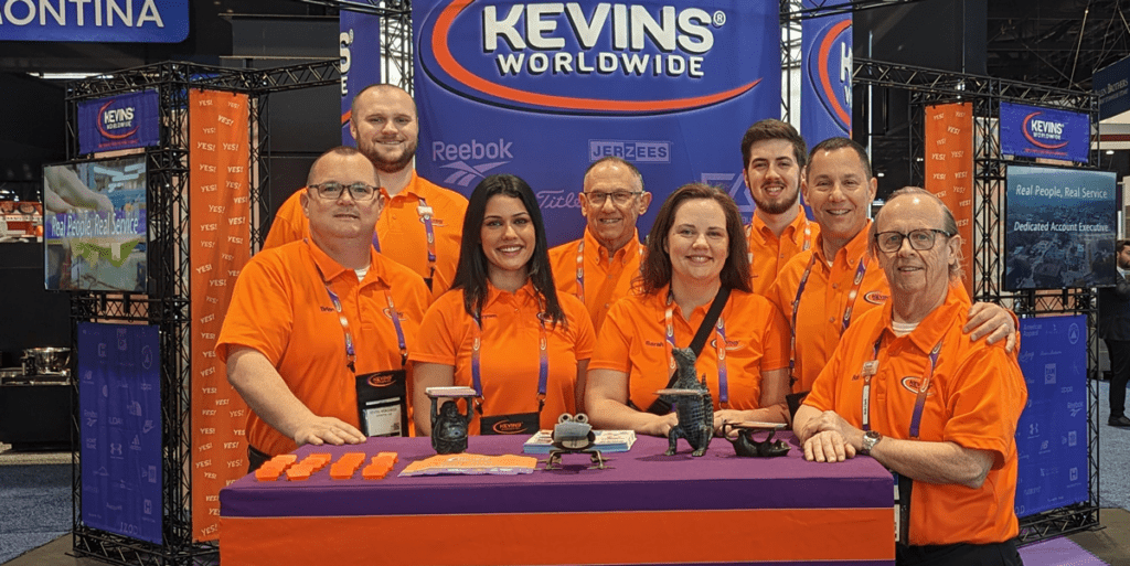 Eight members of the Kevins Worldwide team at a tradeshow booth, all wearing orange branded polos and standind behind a purple table with the Kevins Worldwide logo on it.